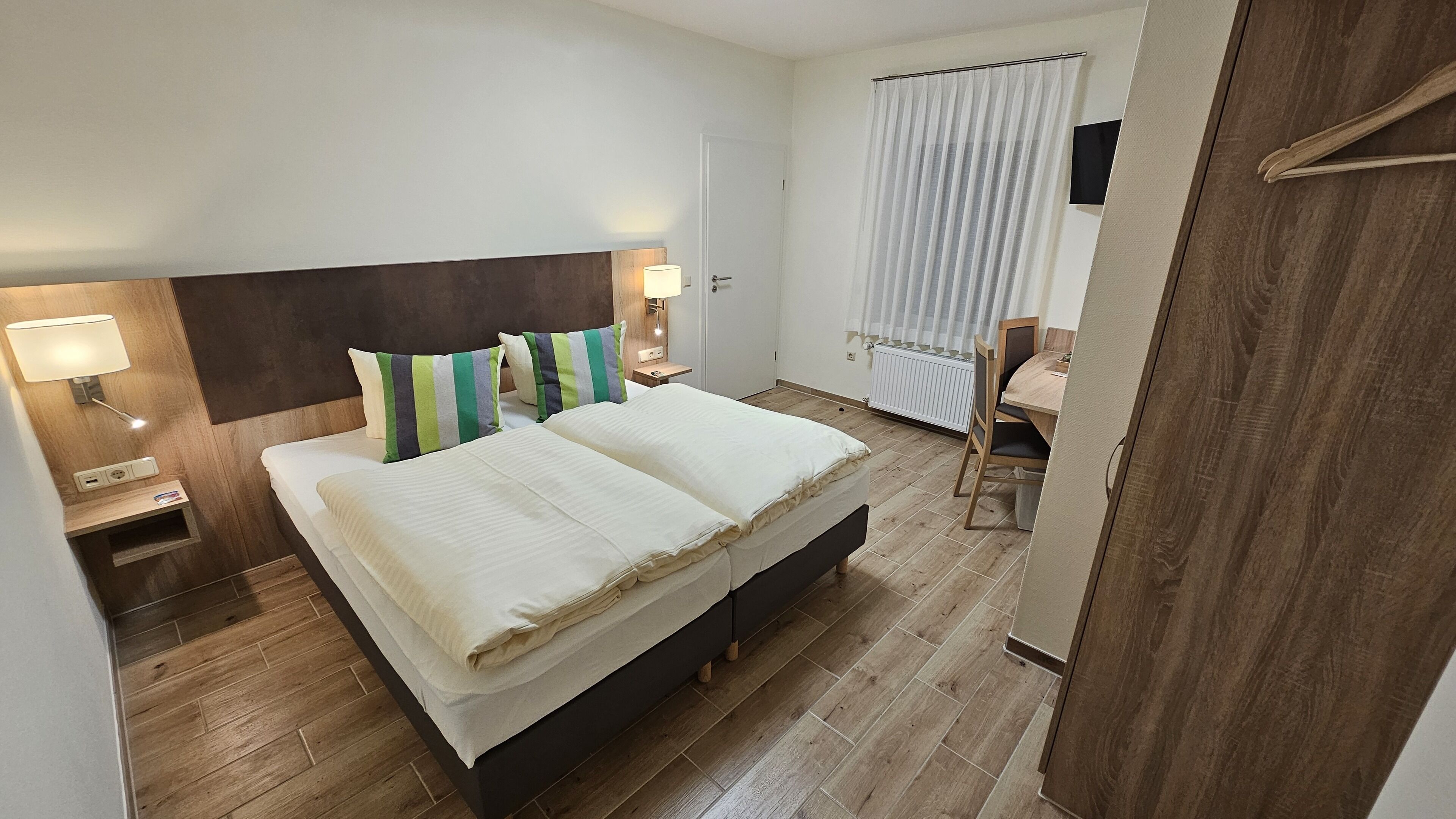 Habitación de hotel con una cama grande, lámparas de noche, cabecero de madera, una mesita y una silla.
