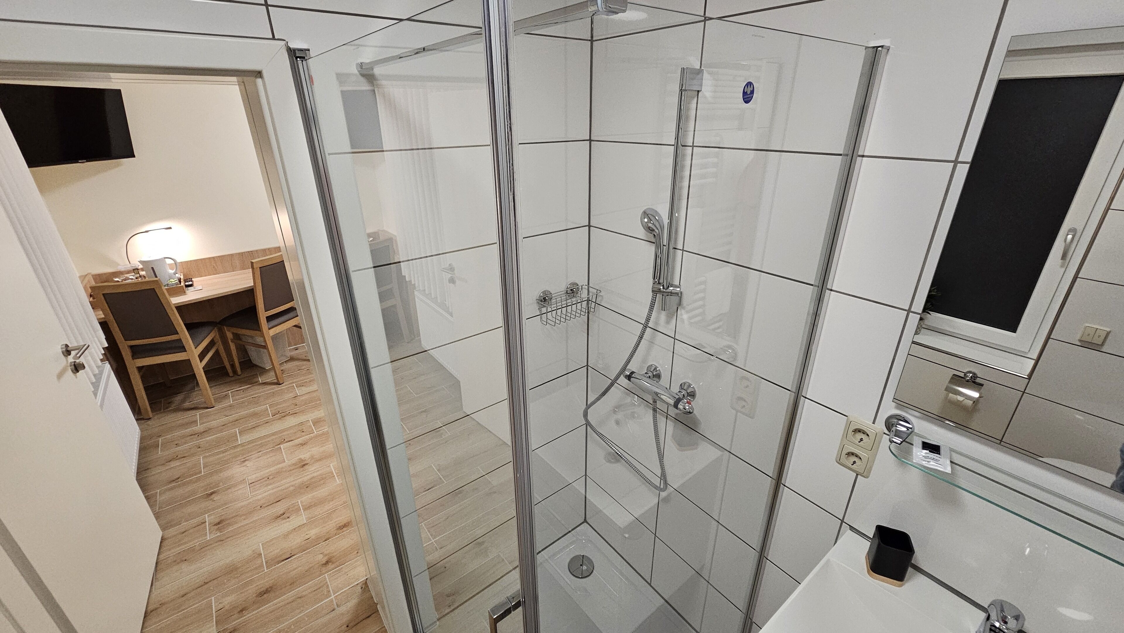 A modern bathroom with a glass shower enclosure, a wooden floor, and a small desk with a chair.