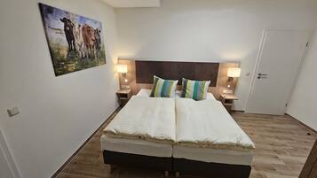 A double bed with white linens, two bedside tables with lamps, a wooden headboard, and a painting of cows on the wall.