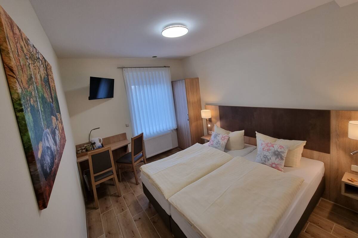 Quarto de hotel com cama, mesas de cabeceira, escrivaninha, cadeira, televisão e janela com cortinas.