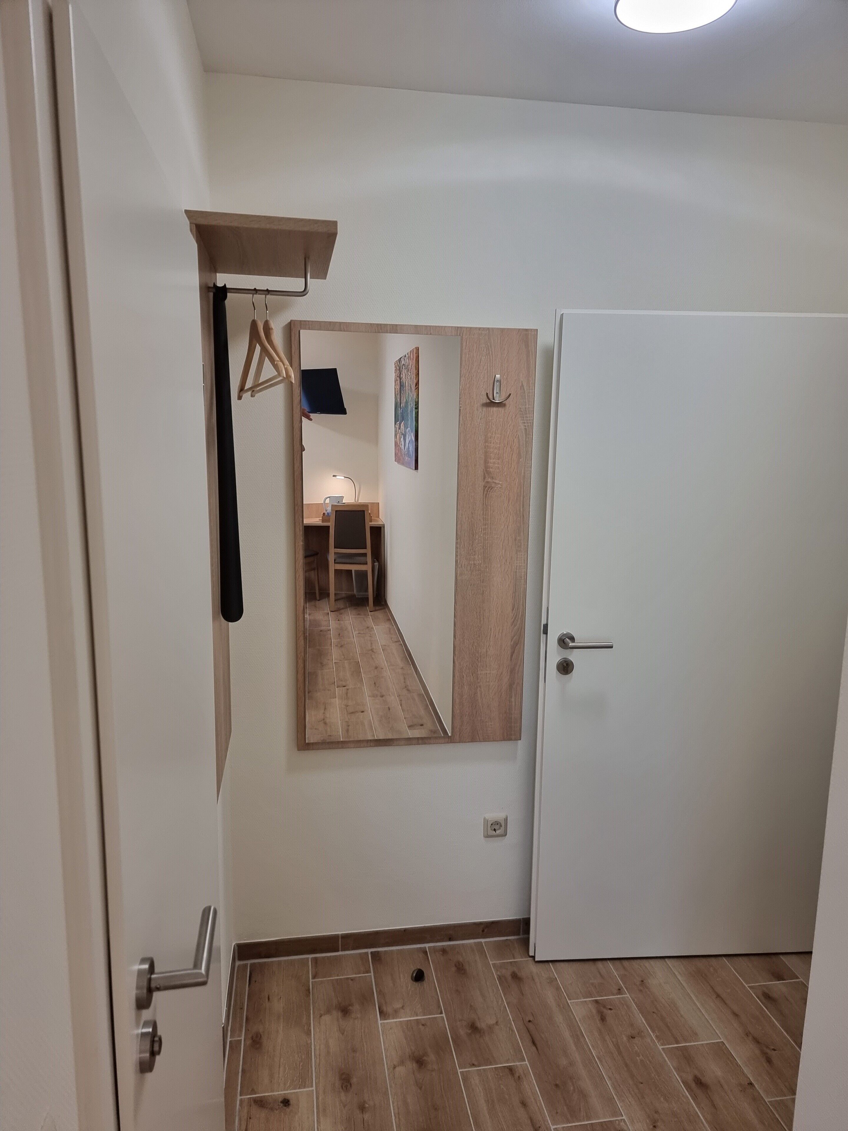 A hallway with a wooden floor, a white door, and a mirror reflecting a room with a desk and chair.