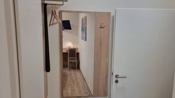 A hallway with a wooden floor, a white door, and a mirror reflecting a room with a desk and chair.