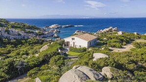 En vit villa med ett tak täckt av tegelpannor, omgiven av frodig grönska och ett kuperat landskap, med utsikt över ett kustnära område och havet i bakgrunden.