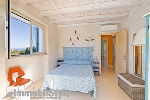 A bedroom with a bed, a chair, a nightstand, a lamp, and a view of the outdoors.