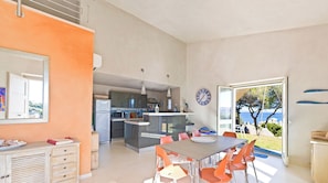 A modern kitchen with a central island, a dining area with orange chairs, and a view of the sea through a large window.