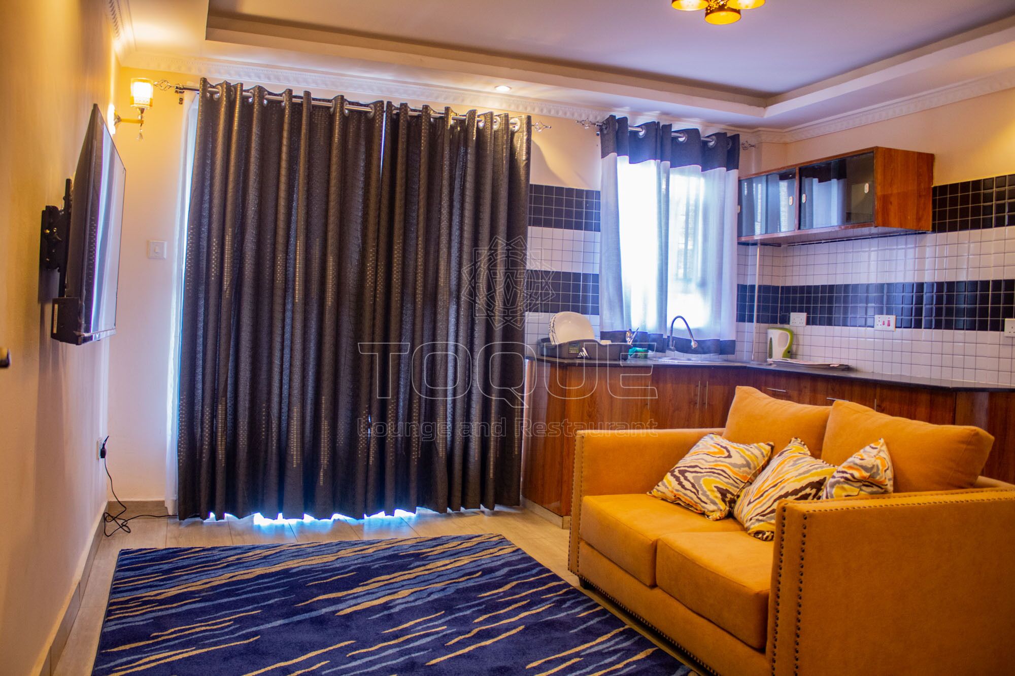 Una sala de estar con sofá, un tapete estampado, una zona de cocina con salpicadero de azulejos y una ventana con cortinas.