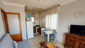 Un espacio habitable compacto con sofá, una pequeña mesa de comedor con sillas, un televisor sobre un mueble de madera y una zona de cocina al fondo.