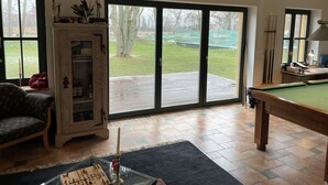 Een woonkamer met een pooltafel, een kast met glazen deuren en een uitzicht op een achtertuin door grote schuifpuien.