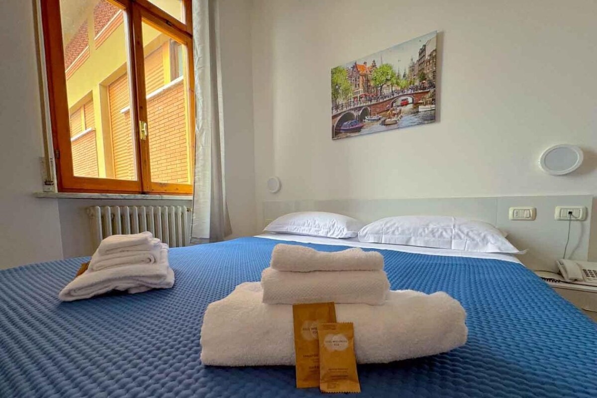 A hotel room with a bed, a painting of a cityscape, a window, and a radiator.