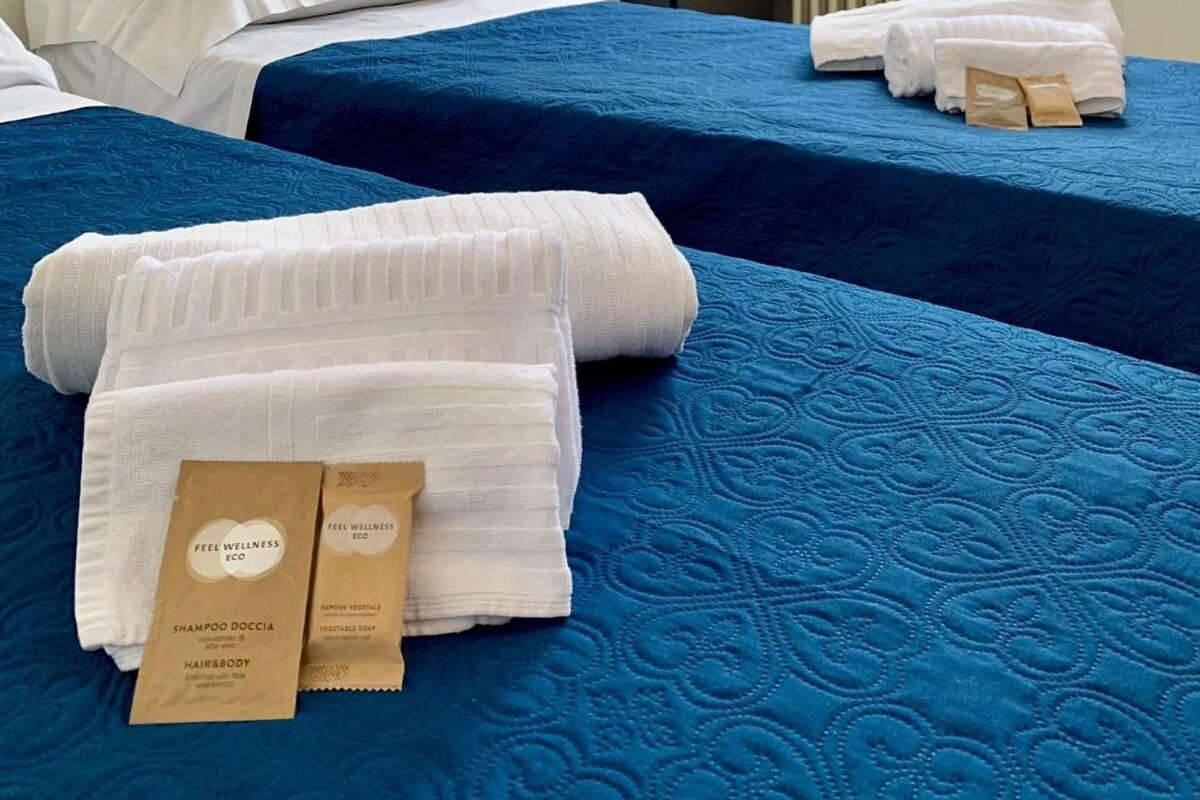 A hotel room with two beds, a radiator, a window with curtains, and toiletries on the beds.