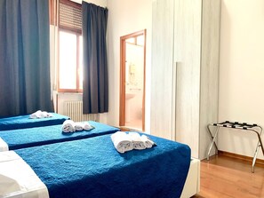 A hotel room with two beds, each covered with blue bedding and white towels. There is a window with dark curtains, a radiator, and a wooden door leading to a bathroom. - Oasi San Francesco (Rome)