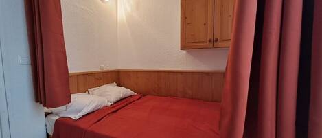 A single bed with a red bedspread, wooden headboard, and a white wall with a light switch and a small wall-mounted light.