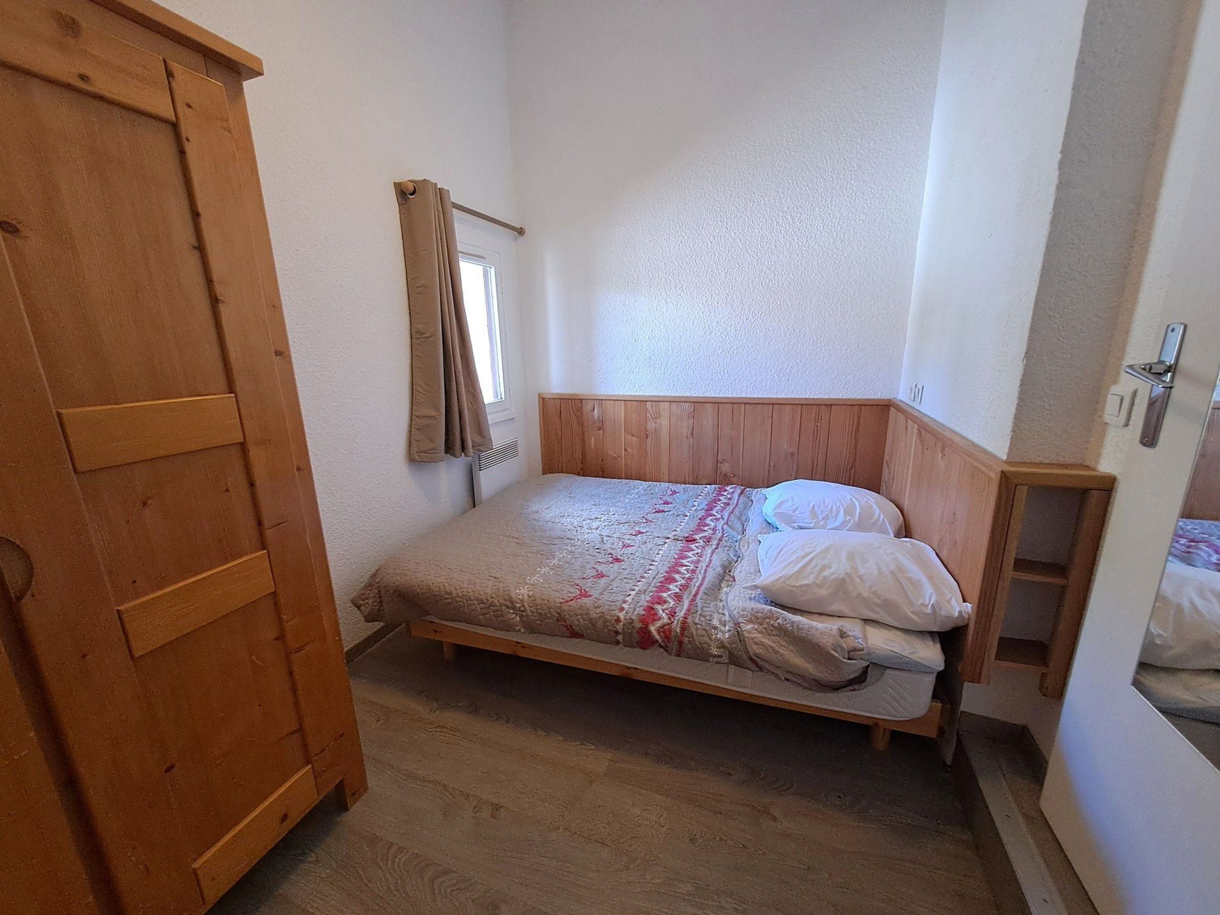 A small, single-bed room with a wooden headboard, a wooden wardrobe, and a window with curtains.