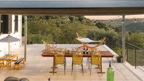 An outdoor dining area with a wooden table set for four, surrounded by wicker chairs, a hammock, and a view of a lush hillside. - Nice villa with pool near Noto, Sicily (Noto)