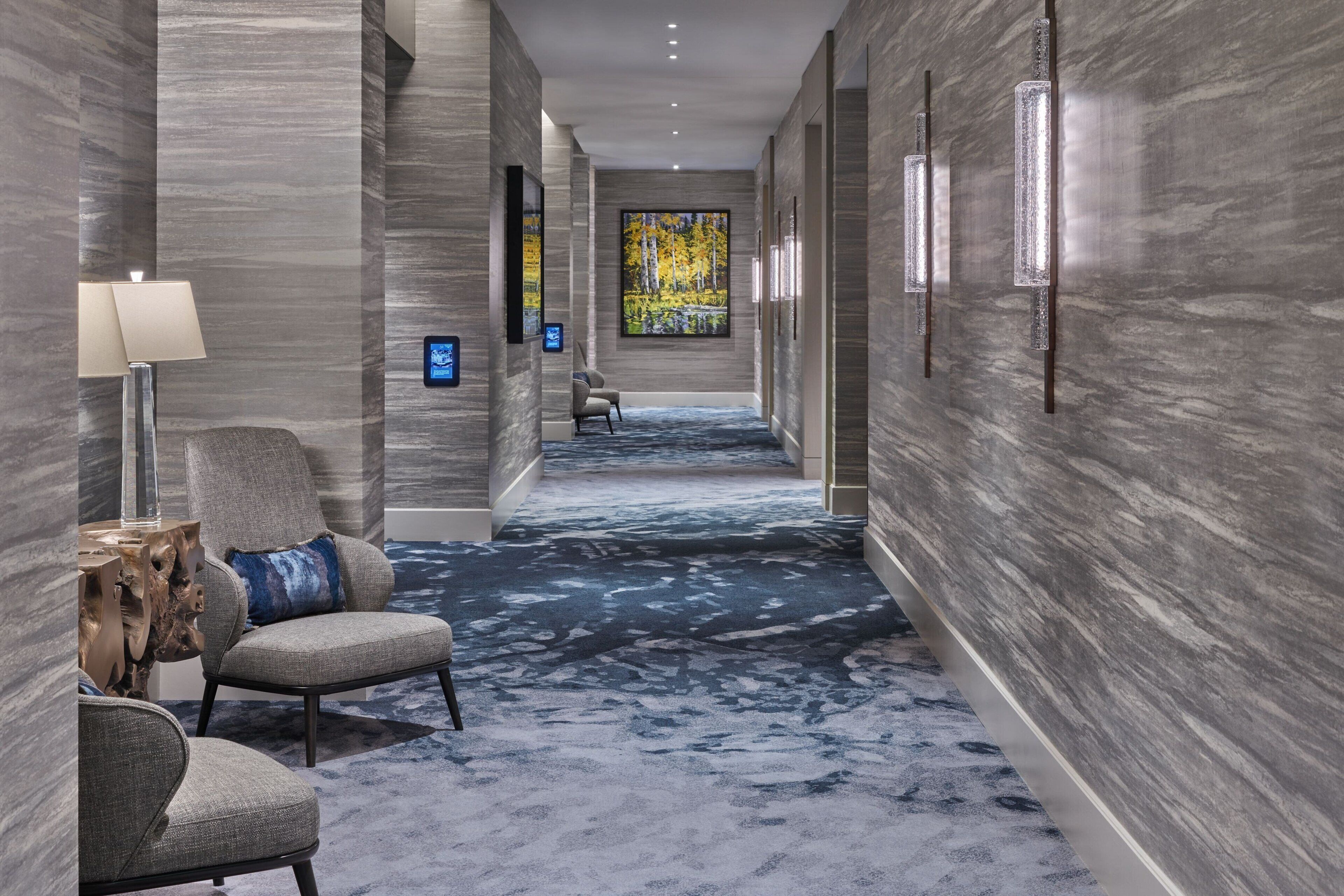 A hallway with a patterned carpet, a chair with a cushion, a side table, and a wall-mounted lamp.