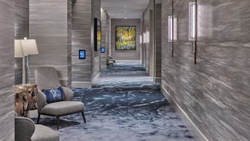 A hallway with a patterned carpet, a chair with a cushion, a side table, and a wall-mounted lamp.