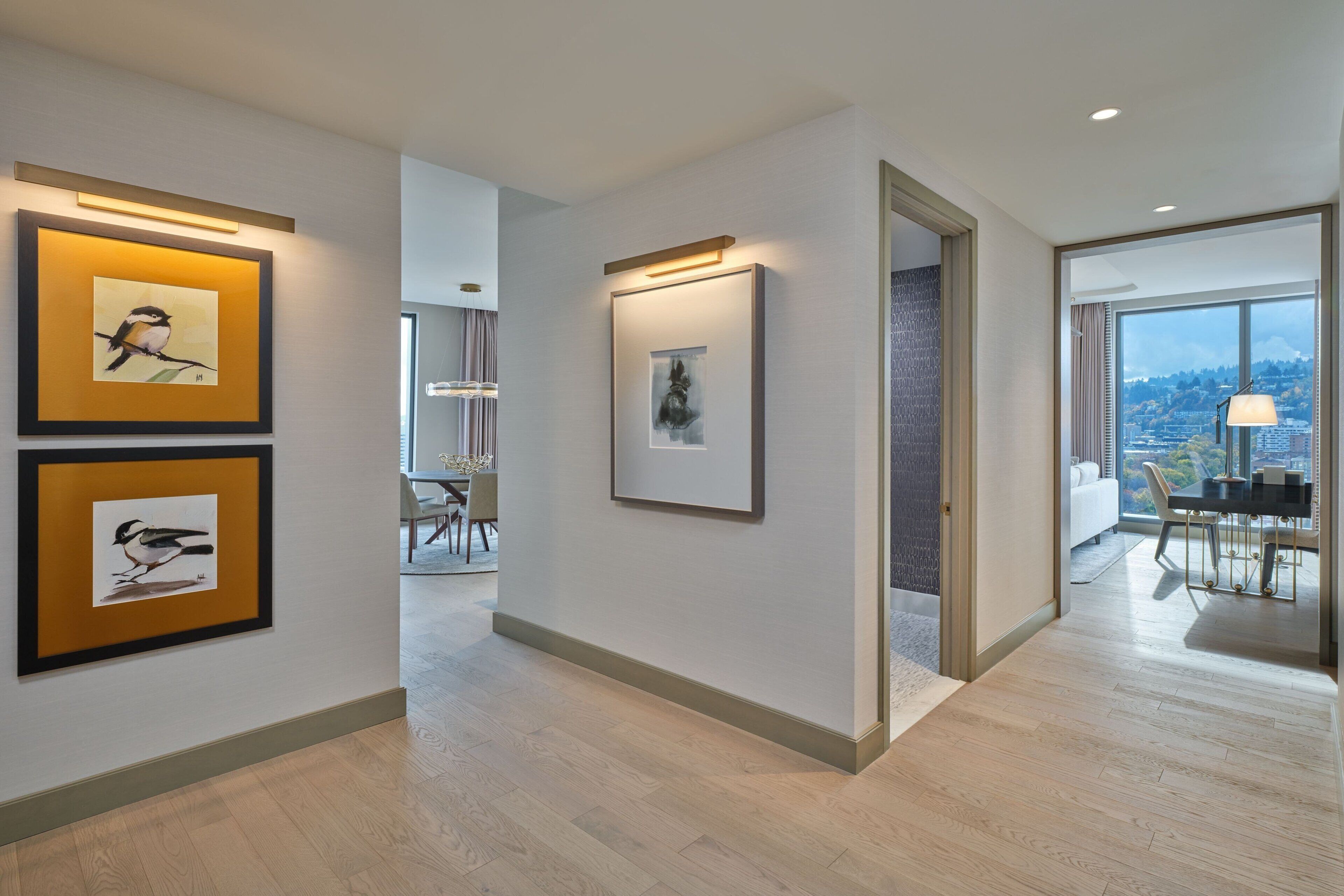 A modern hotel corridor with framed artwork on the walls, a dining area with a view, and a spacious room with a large window.