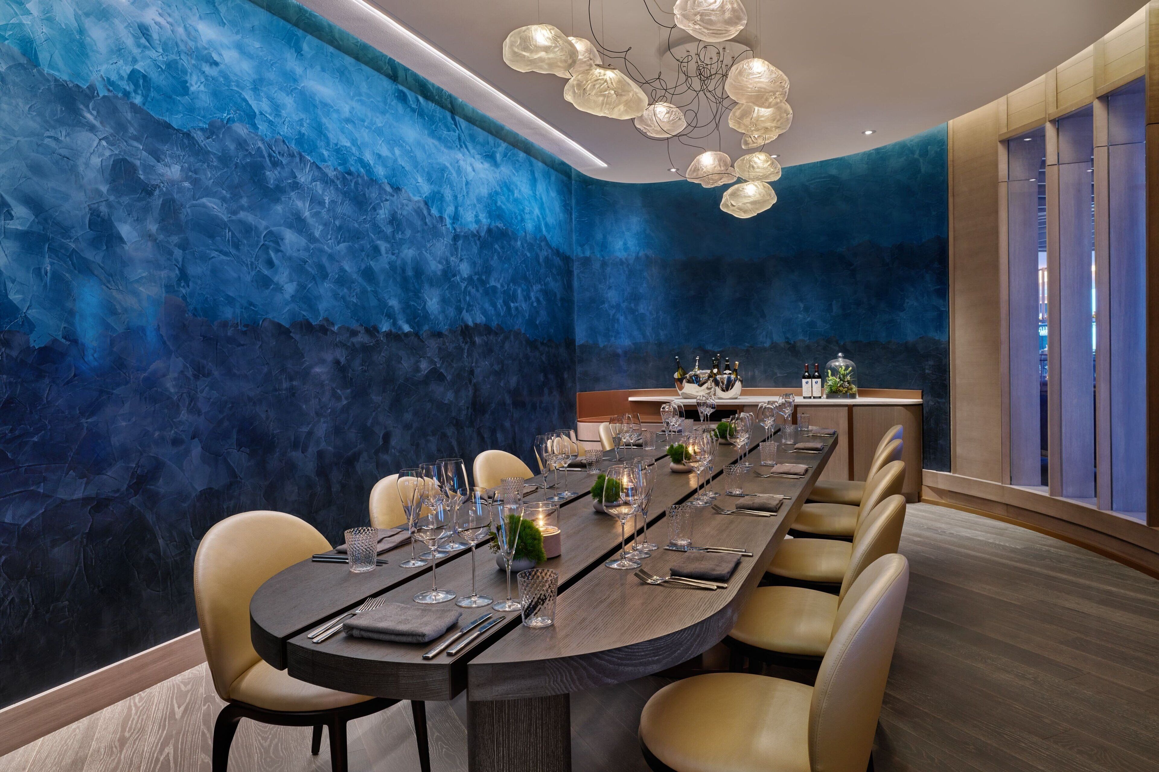 A modern dining room with a long table set for a meal, featuring a blue textured wall and a contemporary chandelier.