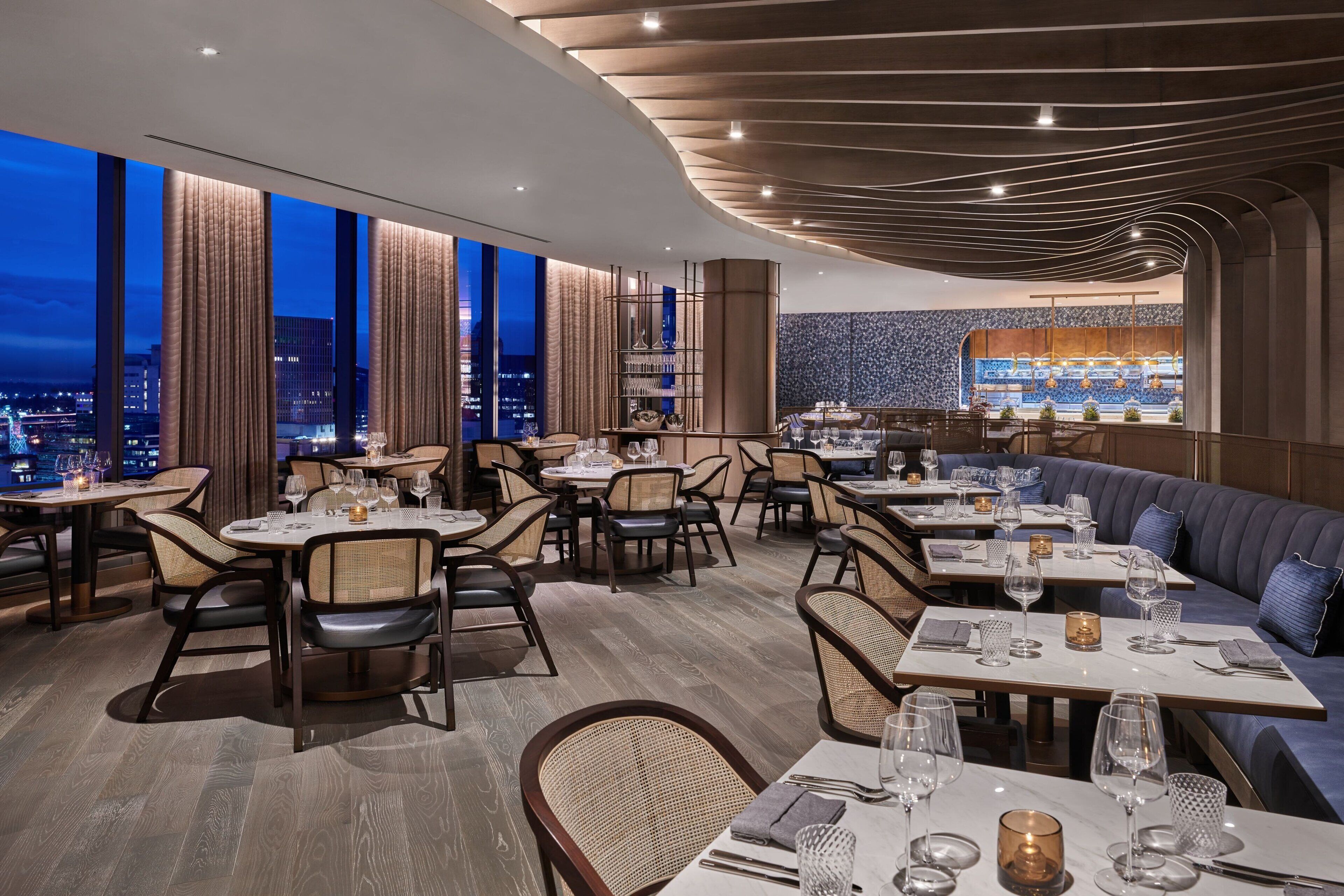 Um restaurante moderno com mesas altas, áreas de cabines, janelas amplas com cortinas e vista para a cidade.