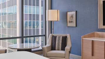 Ein Hotelzimmer mit einem großen Fenster, einem runden Tisch, einem Sessel mit Kissen, einer Stehlampe und einem Schrank.