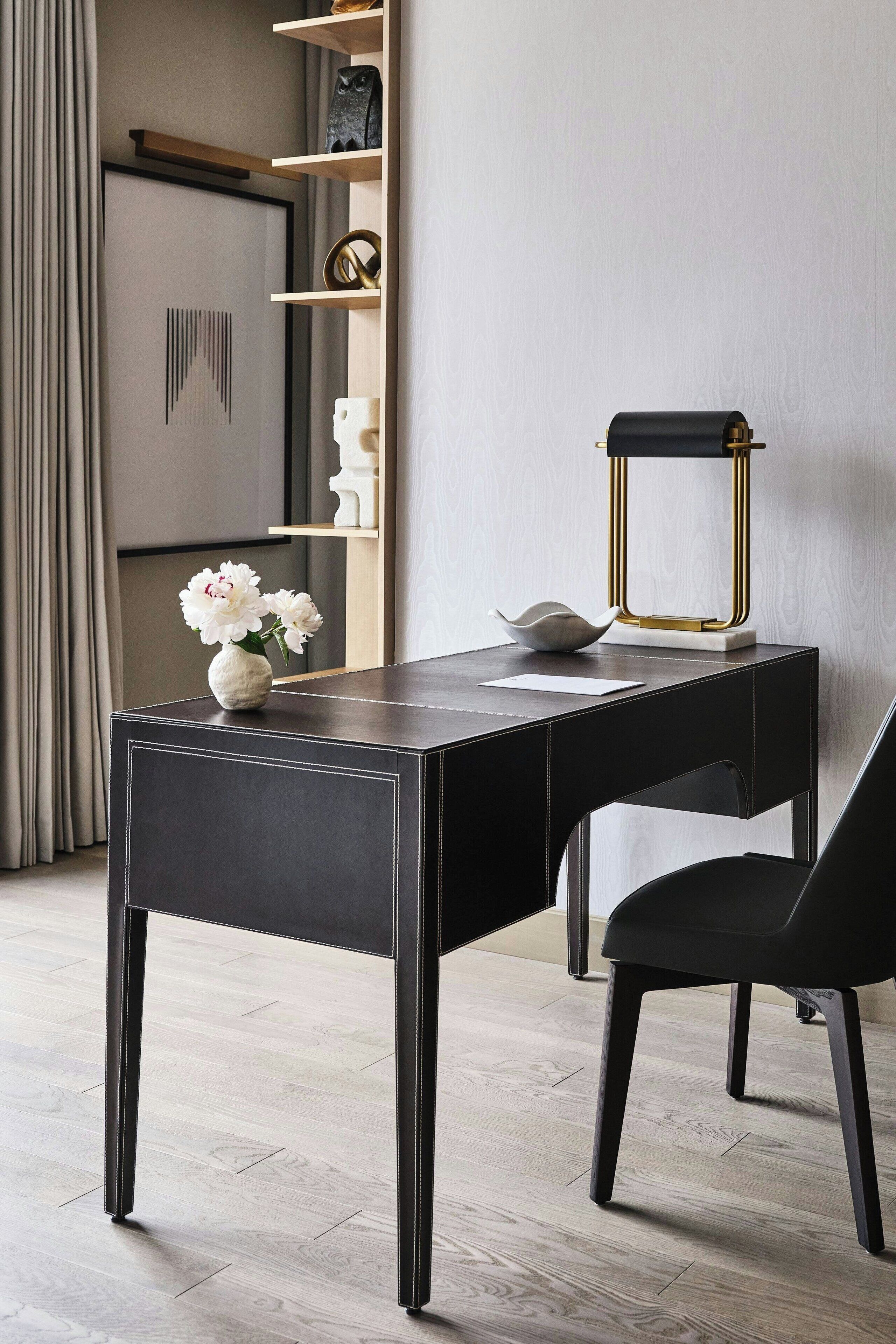 A modern desk with a vase of flowers, a lamp, and a chair in a well-lit room with a wooden floor.