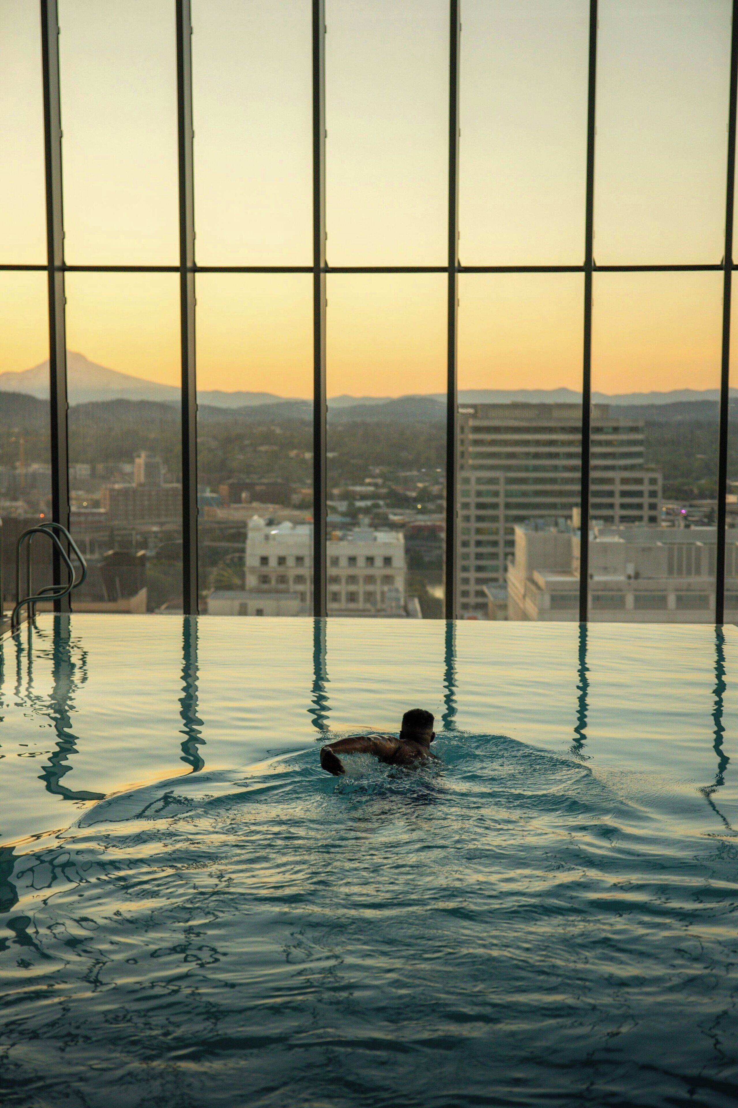 A person swimming in an indoor pool with a cityscape view through large windows.