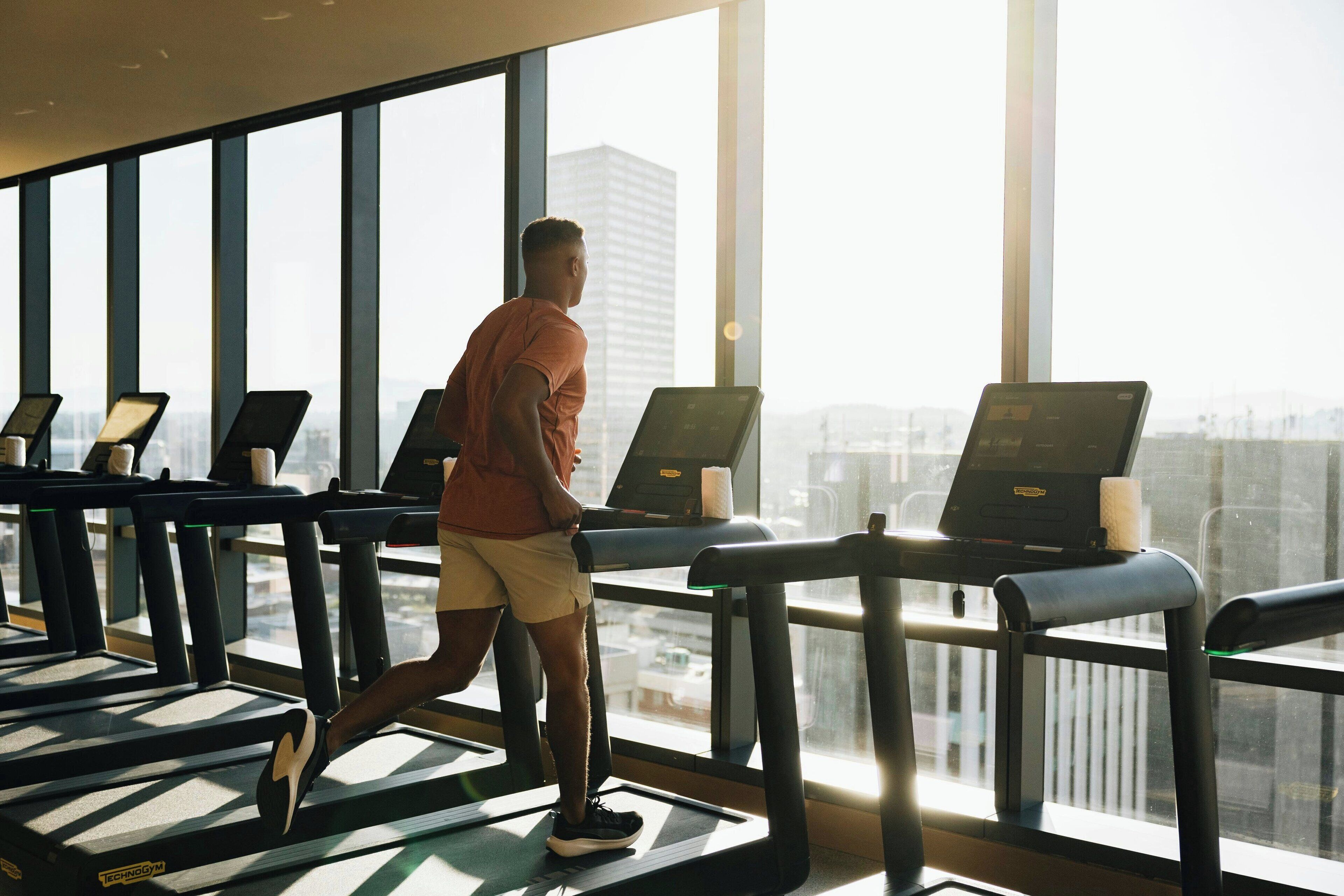 En mand løber på løbebånd i et fitnesscenter med store vinduer og udsigt over byen.
