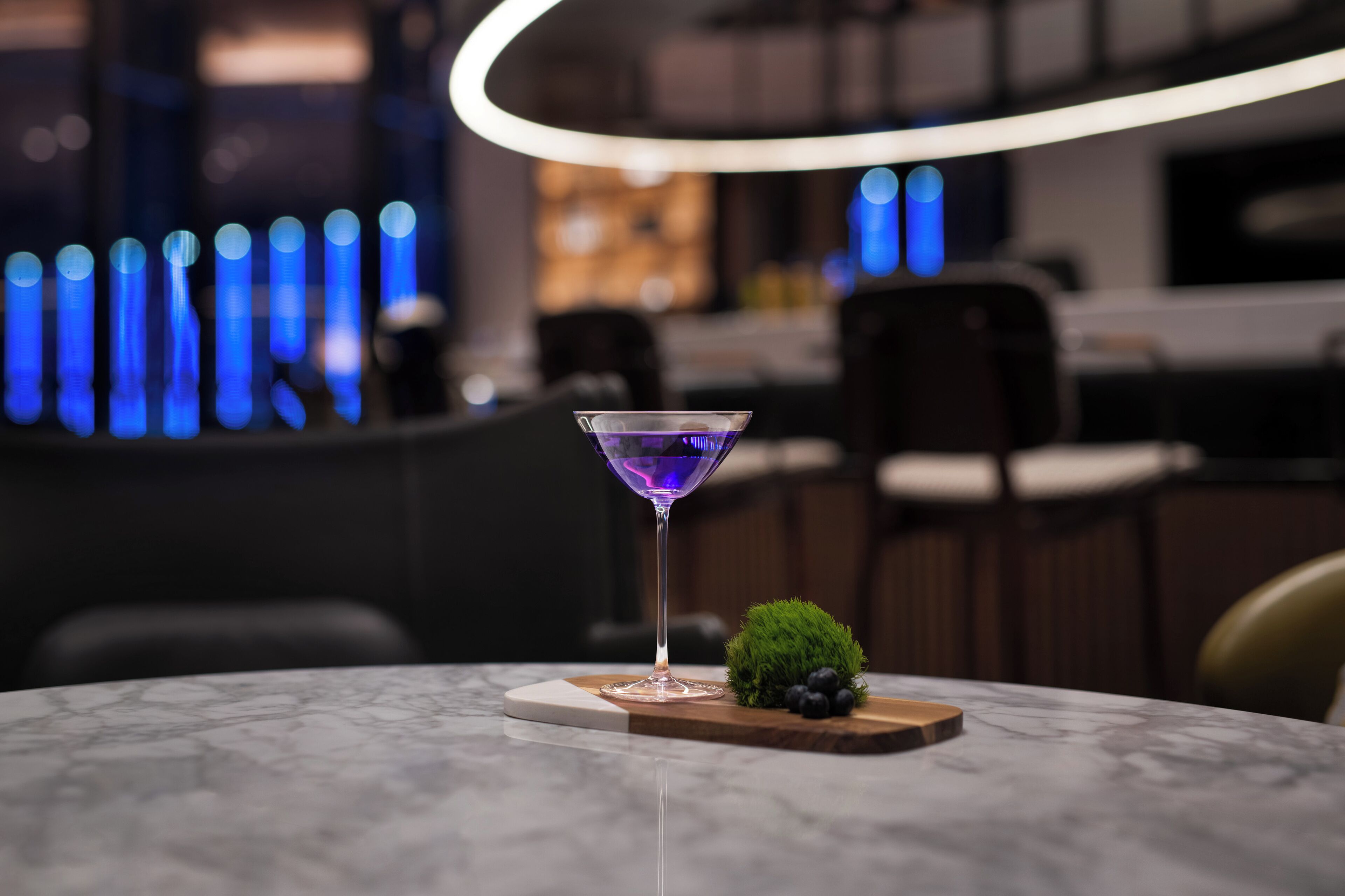 Eine Bar mit Martini-Glas auf einem Marmortisch, einer kleinen grünen Pflanze und einer Holzplatte mit Blaubeeren.