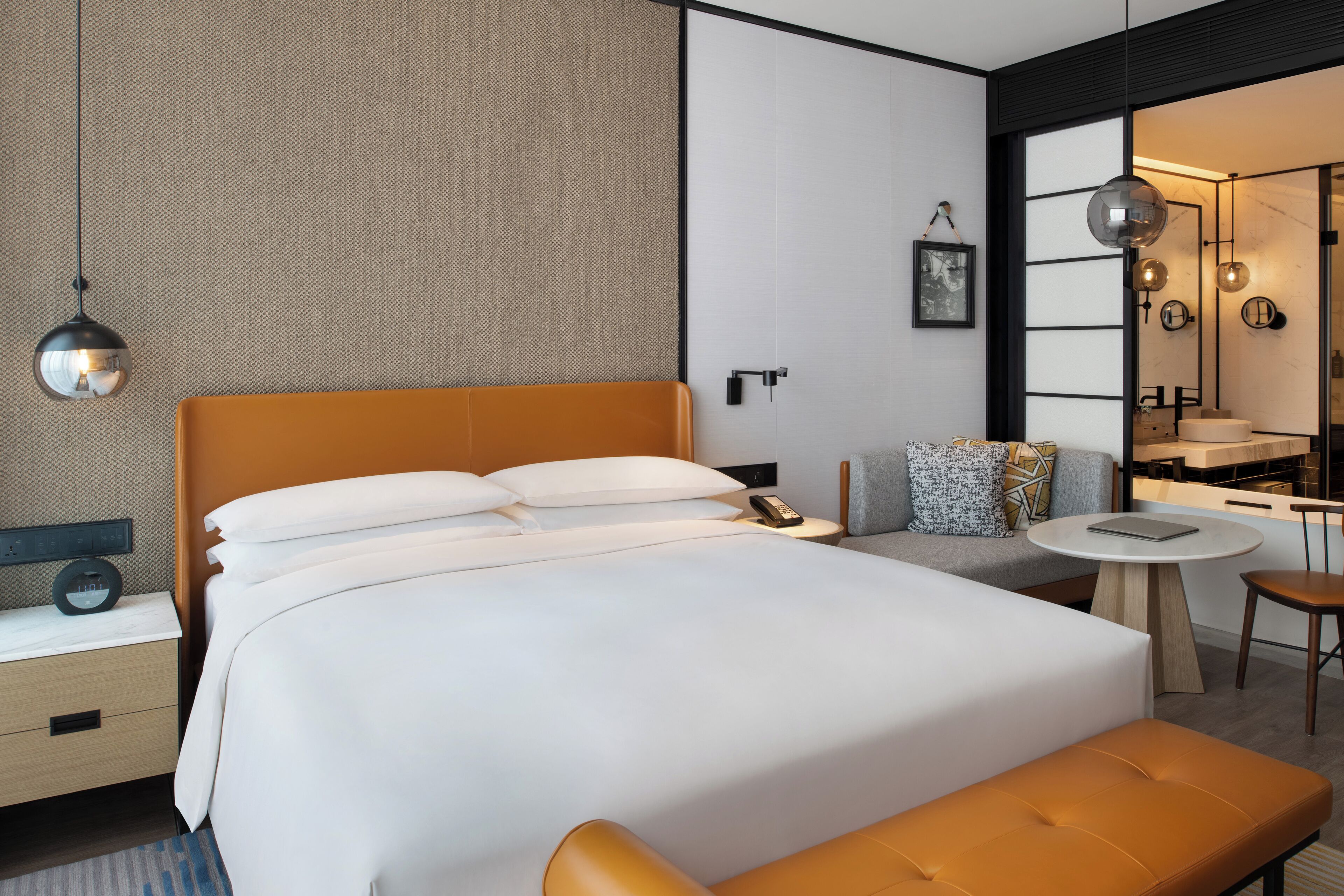 A modern hotel room with a large bed, a bedside table with a lamp, a small round table, and a chair.