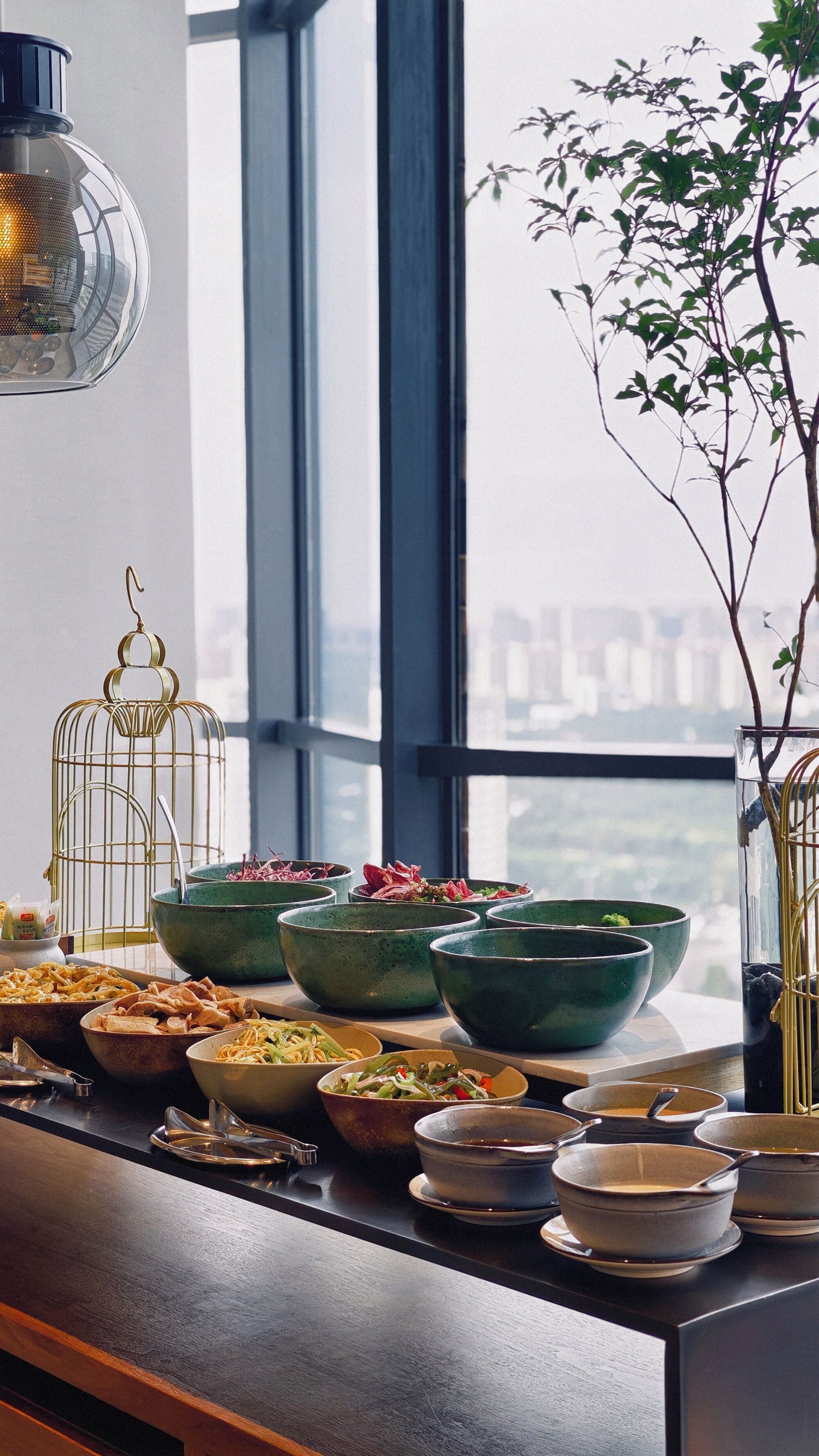 A dining area with multiple bowls and dishes, a hanging light fixture, and a view through large windows.