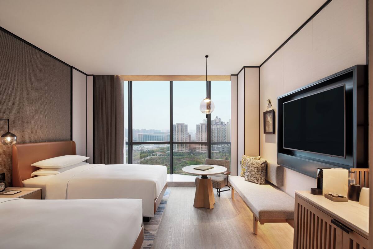 Ein Hotelzimmer mit zwei Betten, einem Fernseher, einem runden Tisch und Blick auf die Stadt.