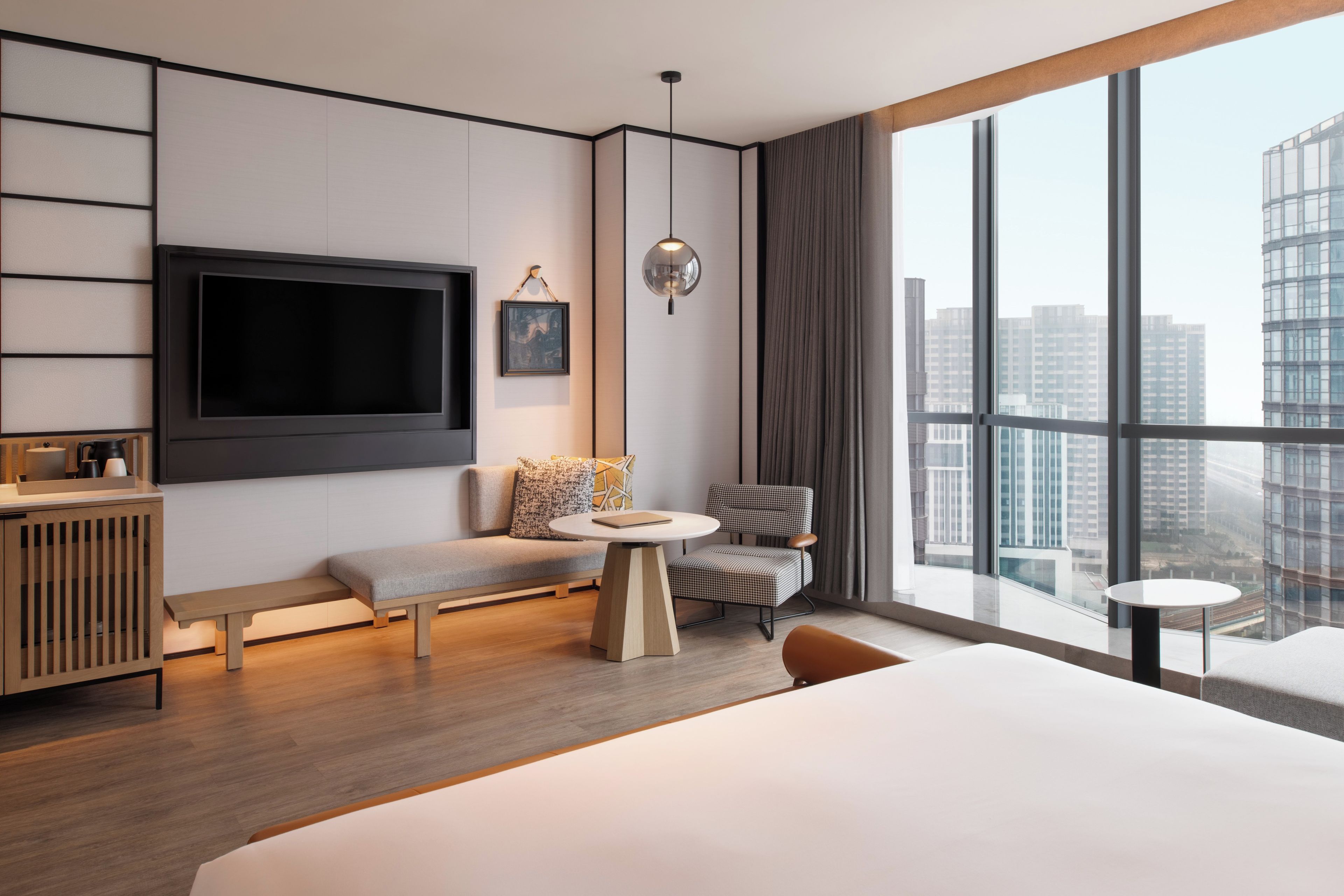 A modern hotel room with a large bed, a seating area with a bench and a chair, a small round table, a TV mounted on the wall, and a large window offering a city view.