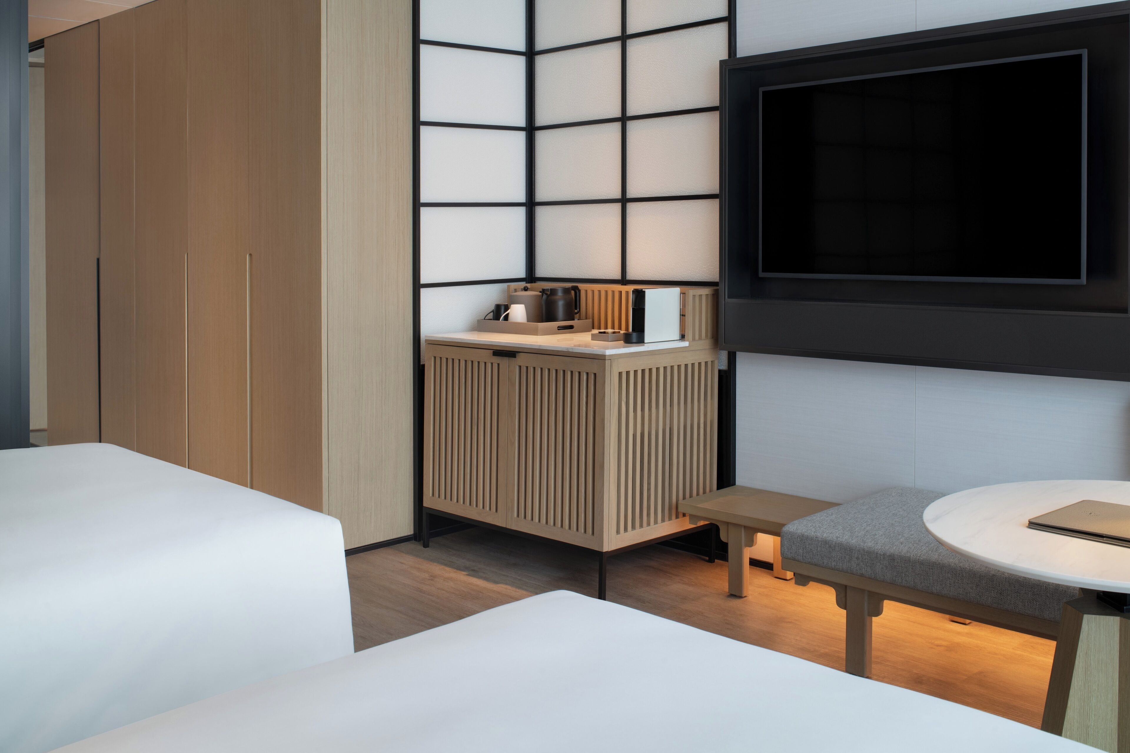 A modern hotel room with two beds, a TV mounted on the wall, a small wooden cabinet, and a round table with a chair.