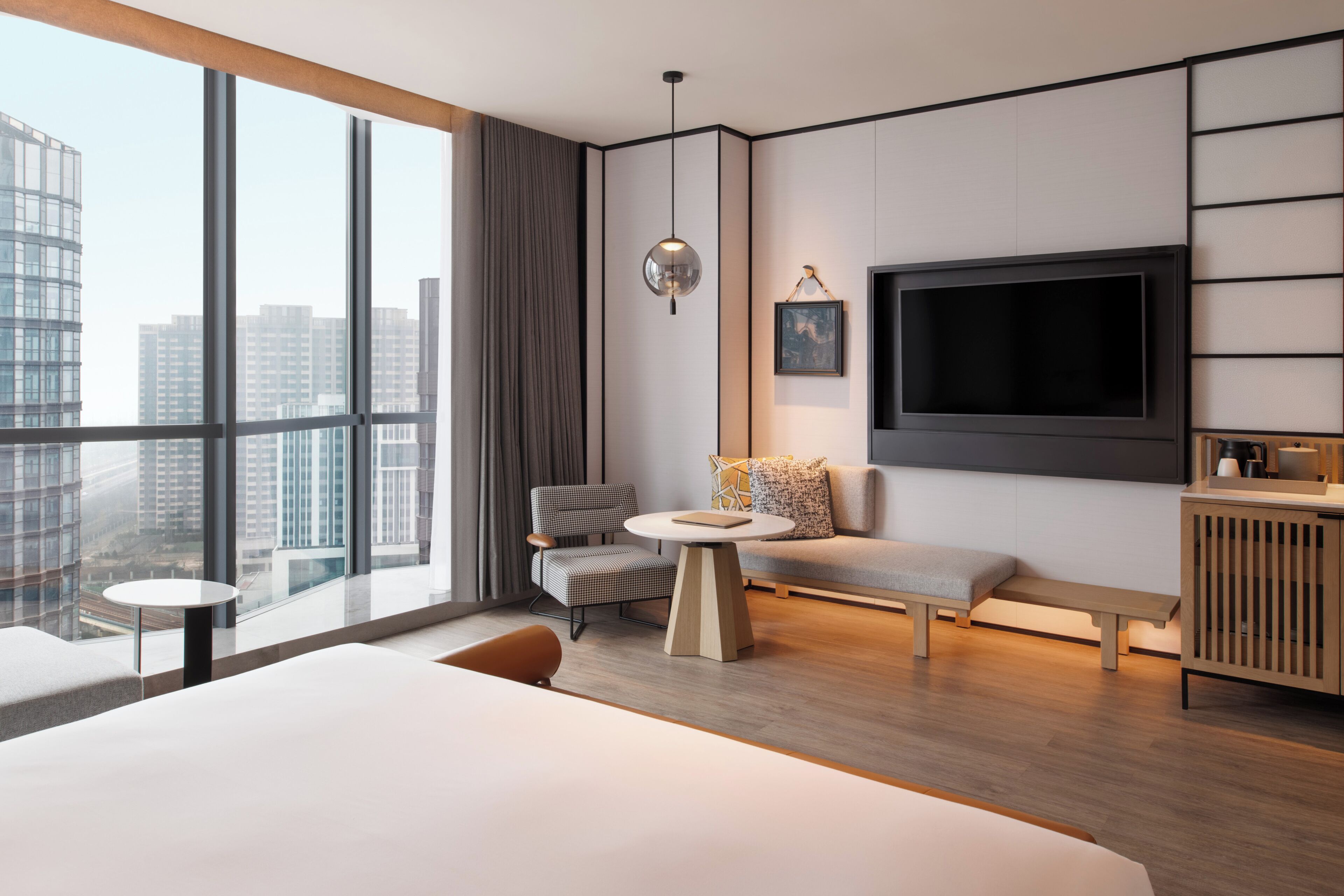 Una habitación de hotel moderna con un ventanal grande, un televisor de pantalla plana, un sofá, una silla, una mesita redonda y un armario de madera.