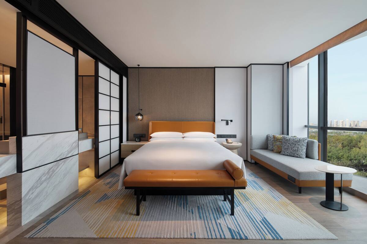 Ein modernes Hotelzimmer mit einem großen Bett, einem Nachttisch, einer Schlaubeleuchtung, einer Couch und einem großen Fenster mit Blick auf die Stadt.