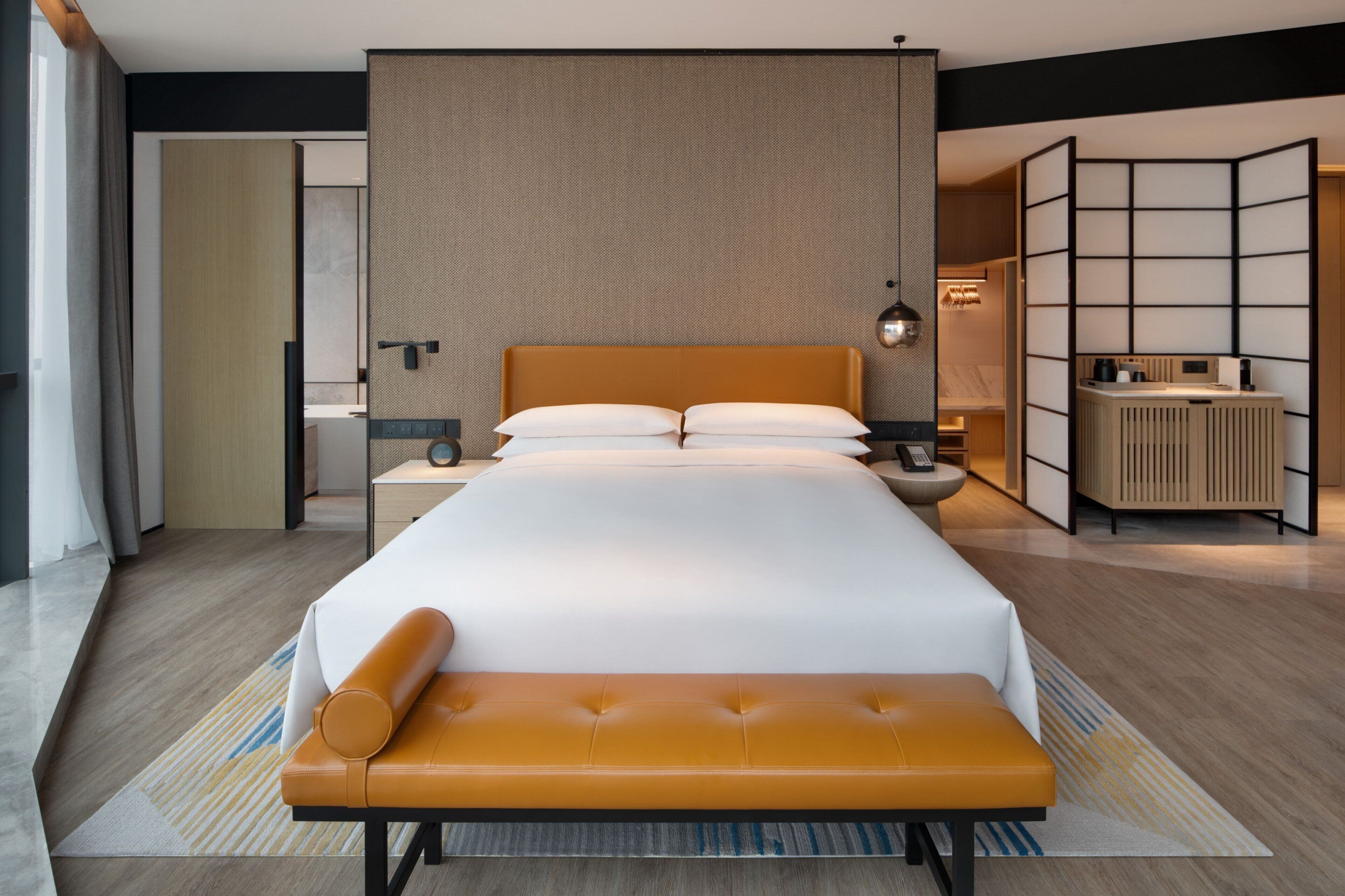 A modern hotel room with a large bed, a bedside table, a bench, and a view of the adjacent room.