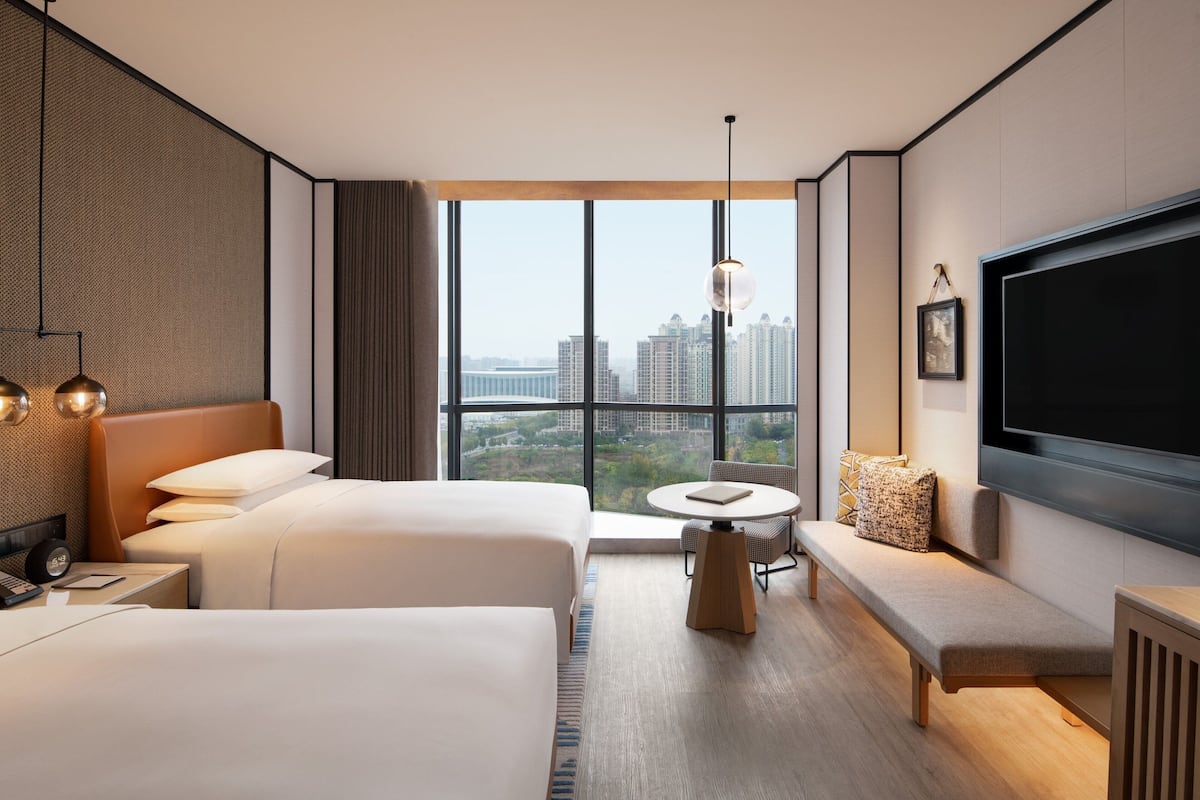 Ein Hotelzimmer mit zwei Betten, einem runden Tisch, einer Bank, einem Fernseher und Blick auf die Stadt.