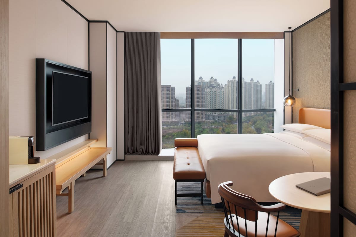 Ein Hotelzimmer mit einem großen Bett, einem Fernseher, einem Schreibtisch mit Stuhl und Blick auf eine Stadtlandschaft.