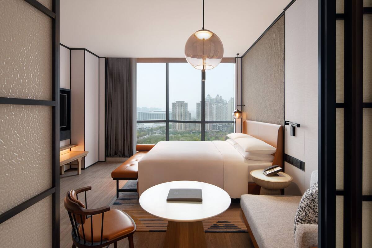 Ein modernes Hotelzimmer mit einem großen Bett, einer Sitzecke mit Tisch und Blick auf die Stadt durch große Fenster.