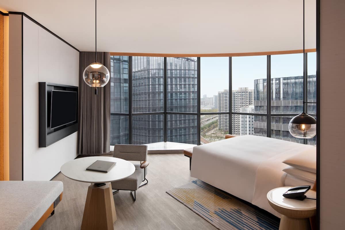 Ein modernes Hotelzimmer mit einem großen Bett, einem Schreibtisch, einem Sessel, einem Fernseher und Blick auf die Stadt durch große Fenster.