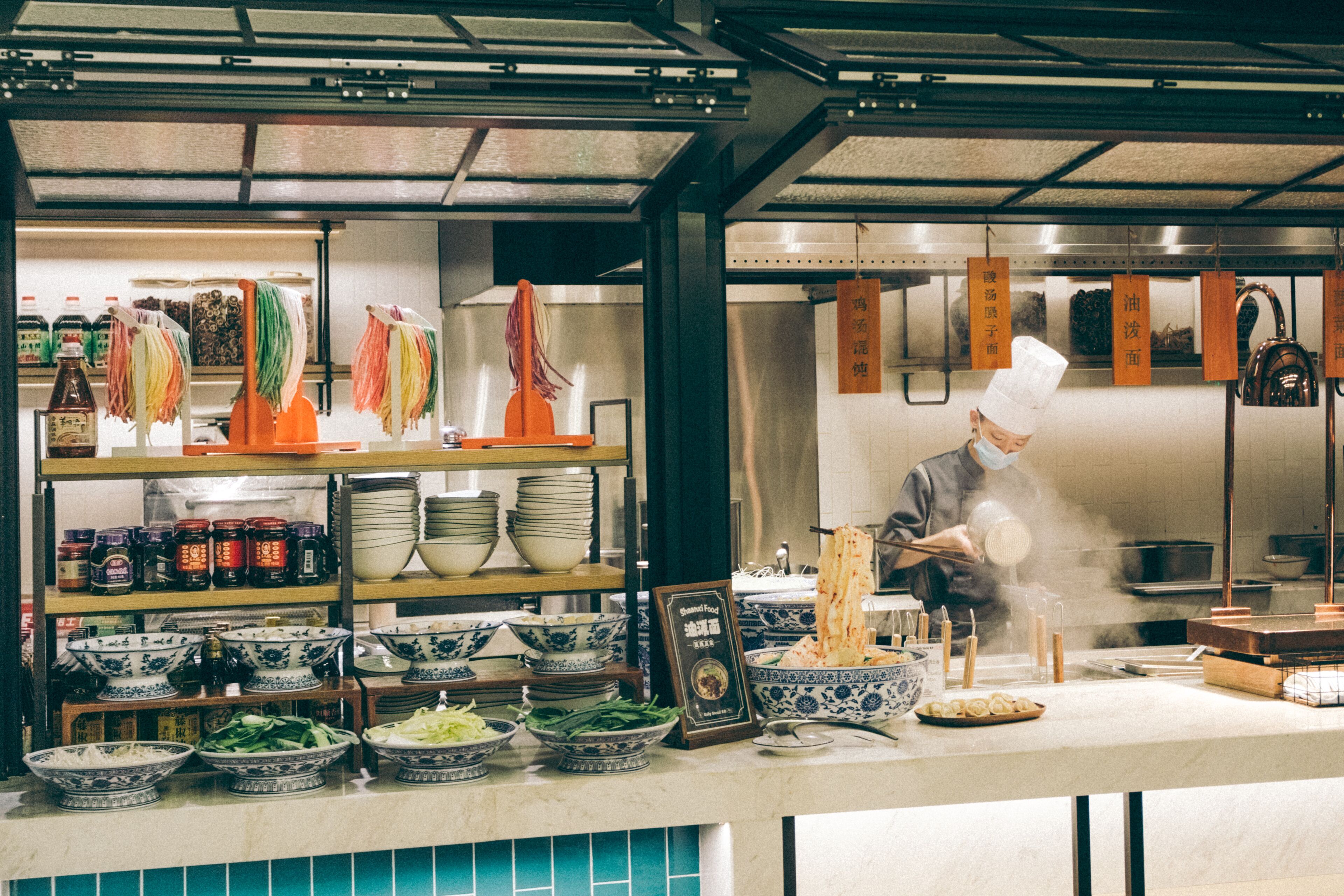 Une cuisine où un chef prépare des plats, des étagères remplies de bols et de condiments, et des ustensiles suspendus.