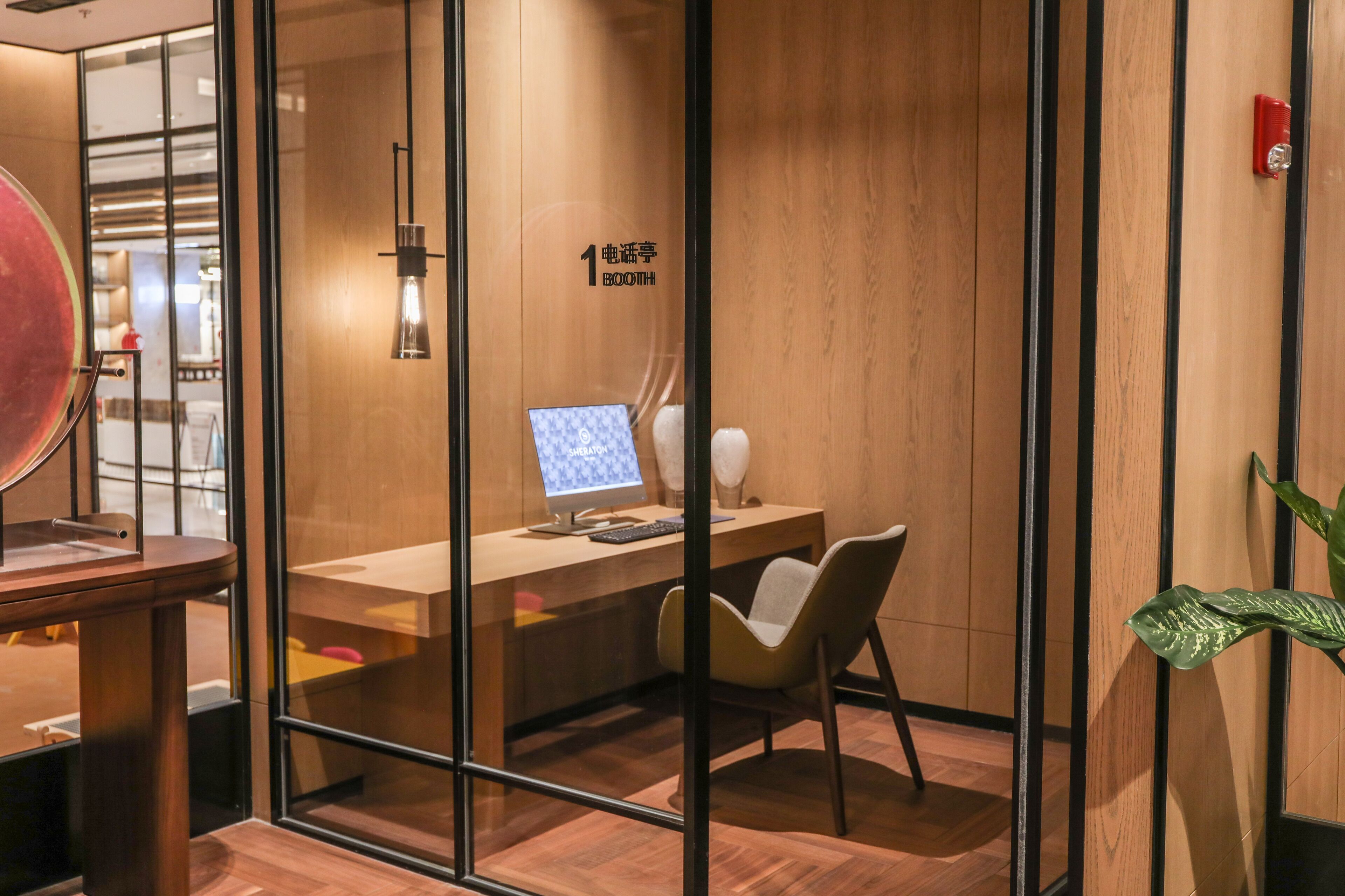 A modern office space with a glass partition, a wooden desk with a computer, a chair, and a potted plant.