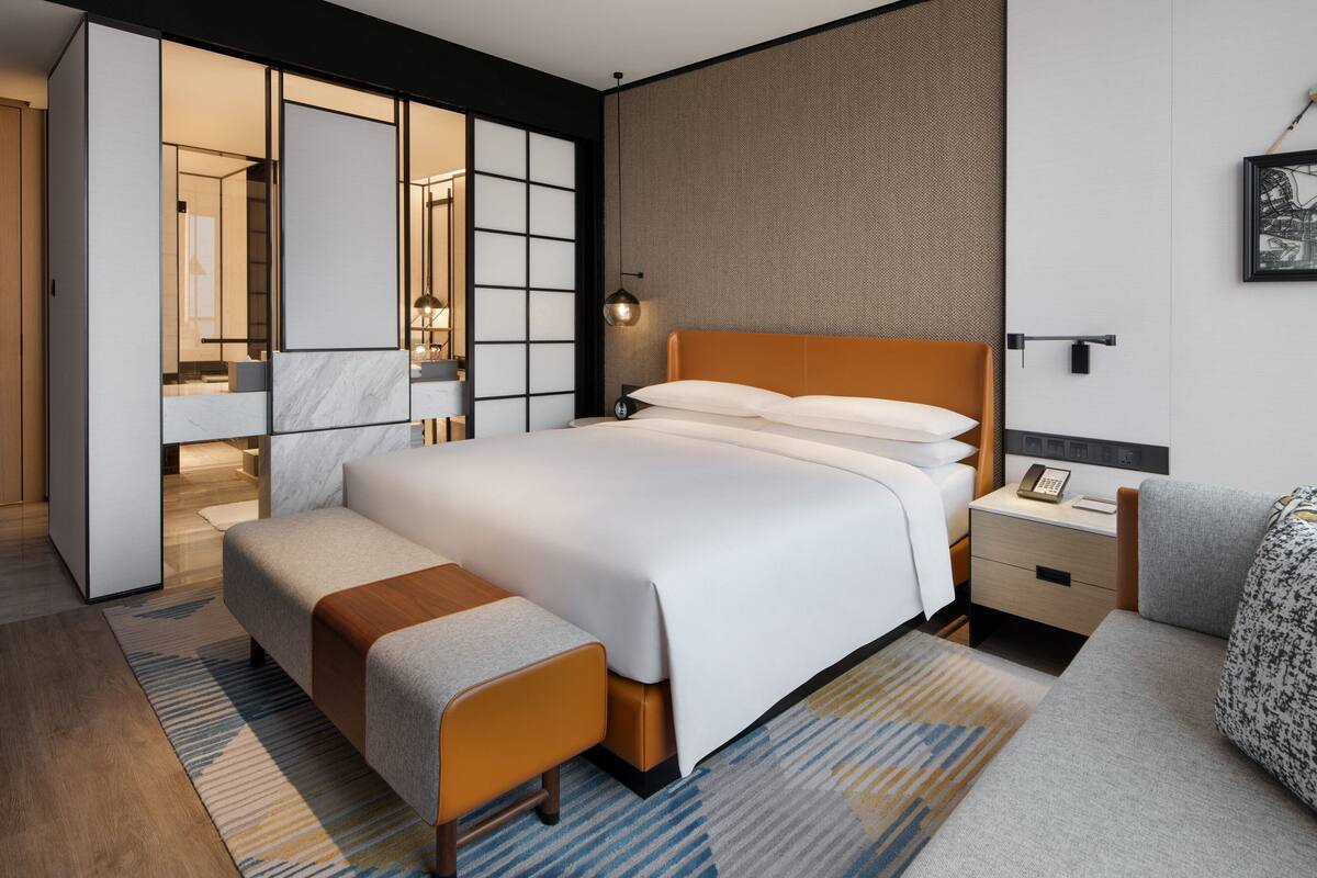 Ein modernes Hotelzimmer mit einem großen Bett, einem Nachttisch mit Lampe, einem kleinen Schreibtisch mit Telefon und einer Sitzecke mit einer Bank.