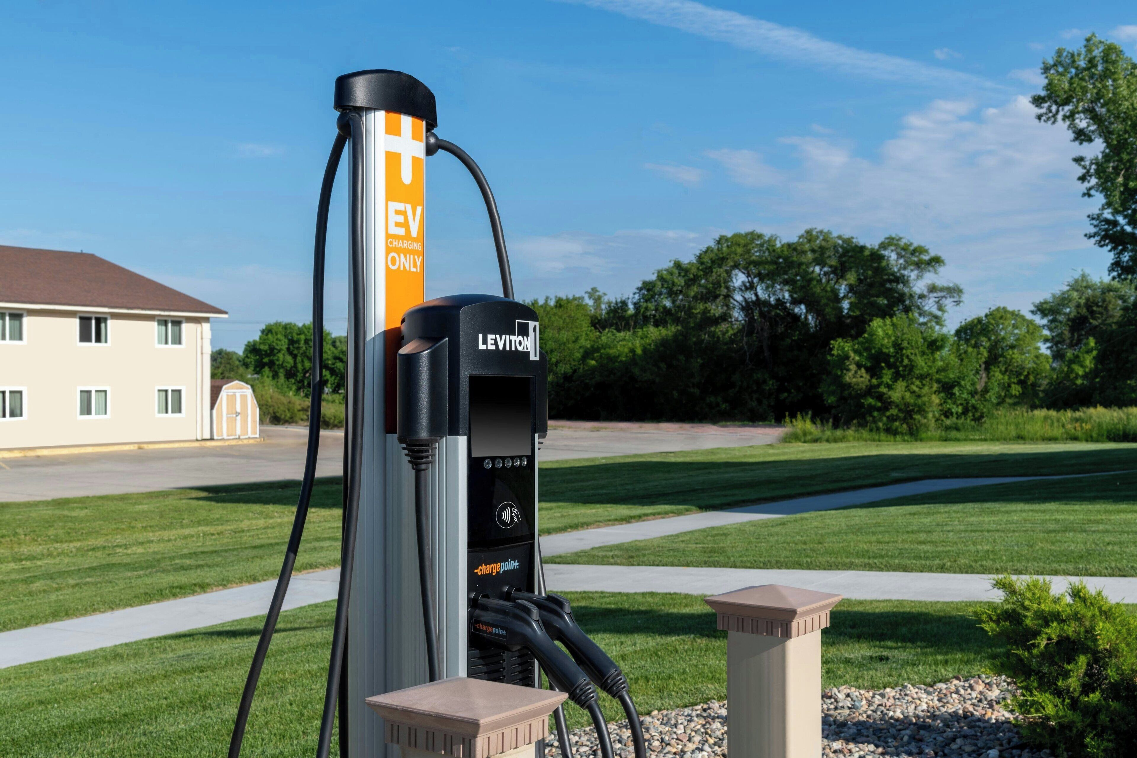 An EV charging station branded with 'LEVITON' and 'EV CHARGING ONLY' in a suburban setting with a house and greenery in the background.