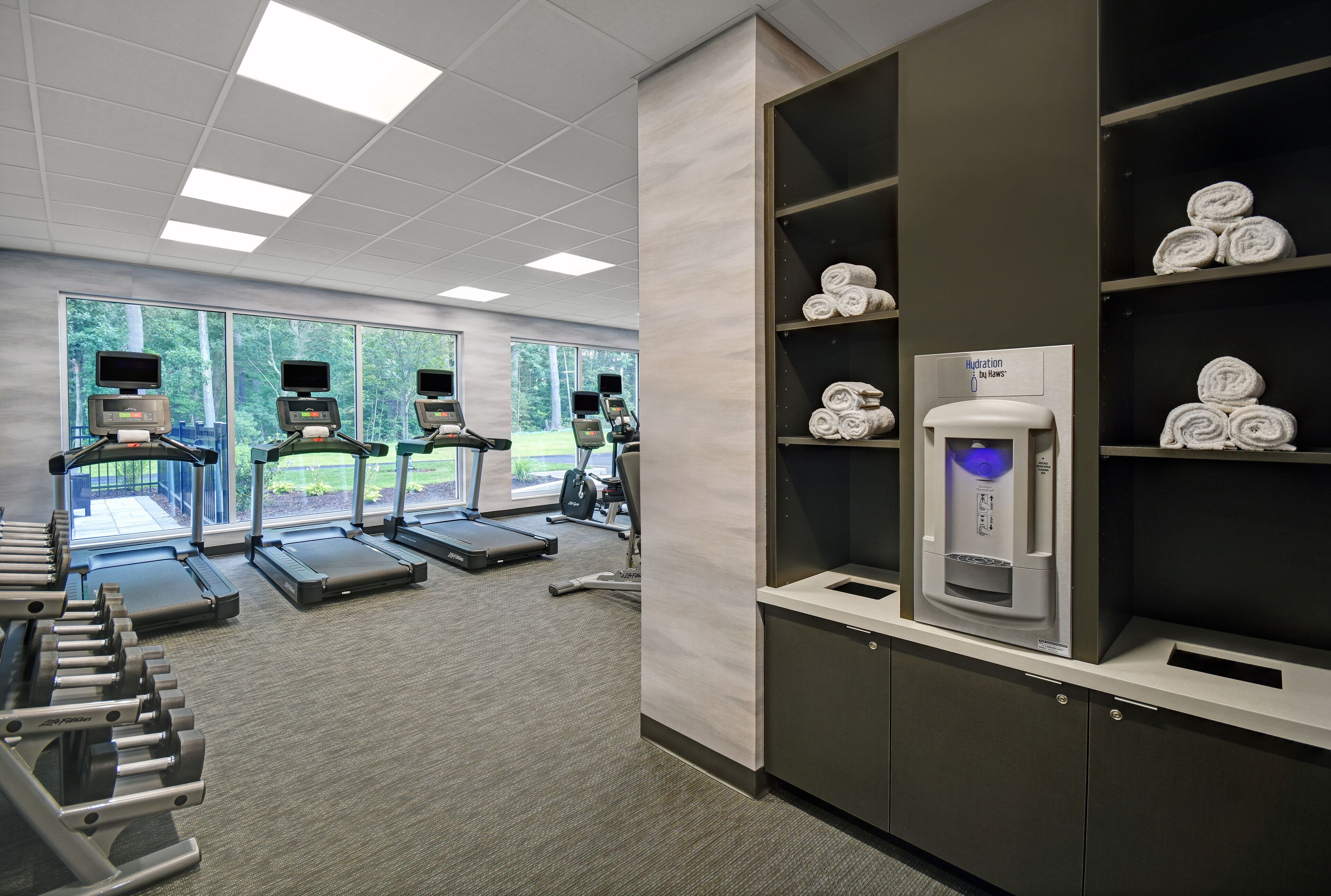A gym with treadmills, ellipticals, and weight machines. There are windows with a view of greenery, and a hydration station with towels on shelves.