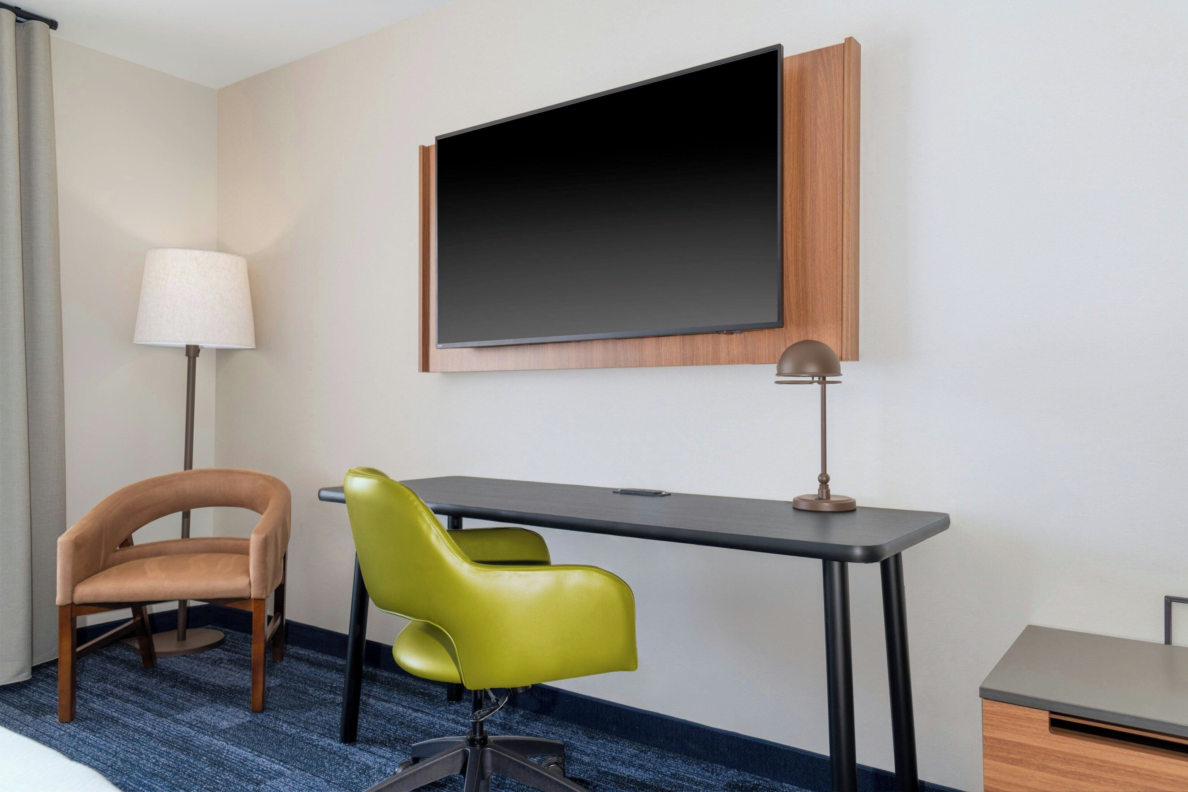 A hotel room with a desk, two chairs, a lamp, and a large flat-screen TV mounted on the wall.