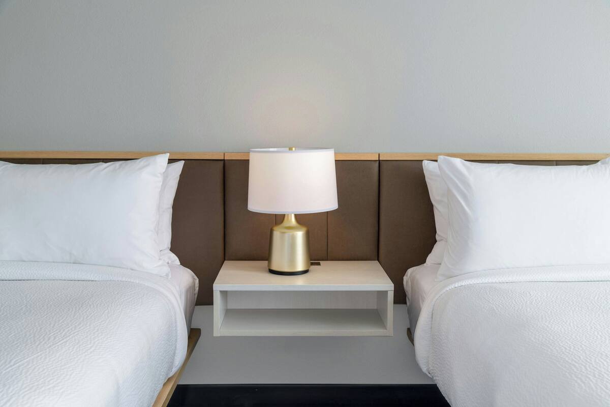 Una habitación de hotel con dos camas, una mesita de noche con lámpara y una pared lisa.