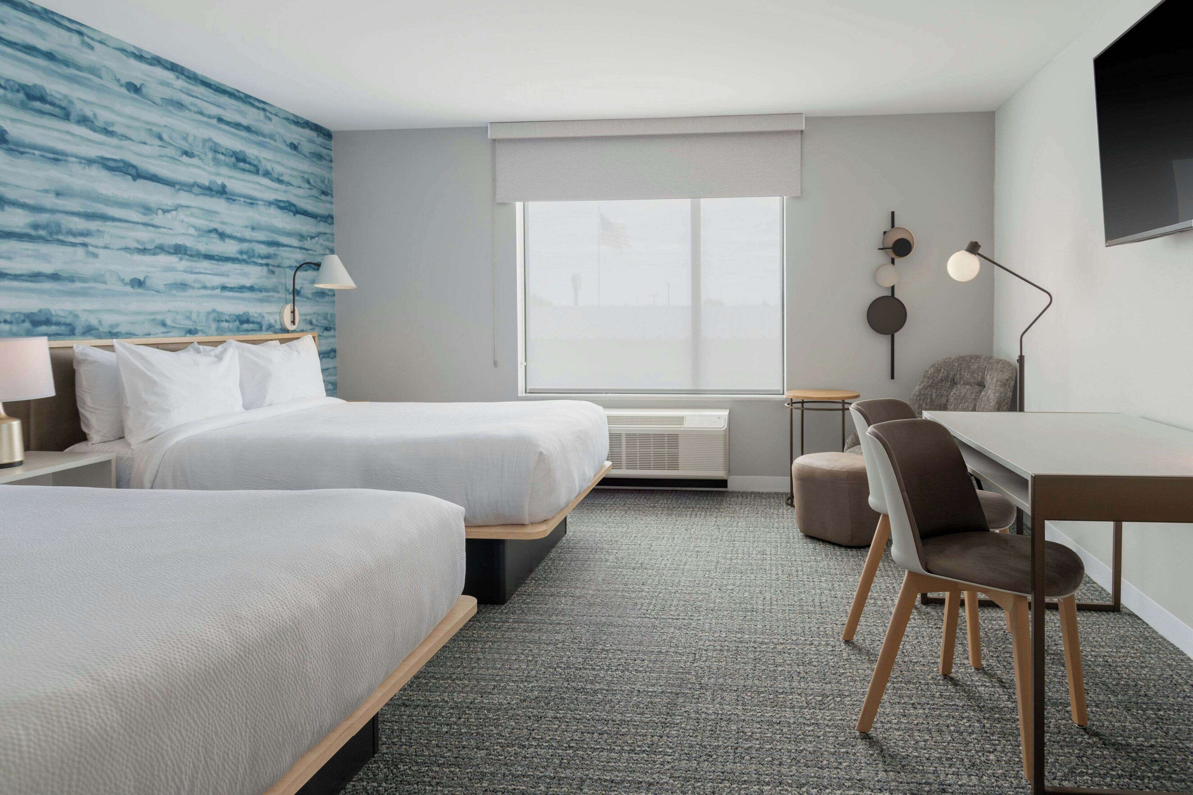 A hotel room with two beds, a desk, and a chair. There is a large window, a wall-mounted TV, and a wall art piece.