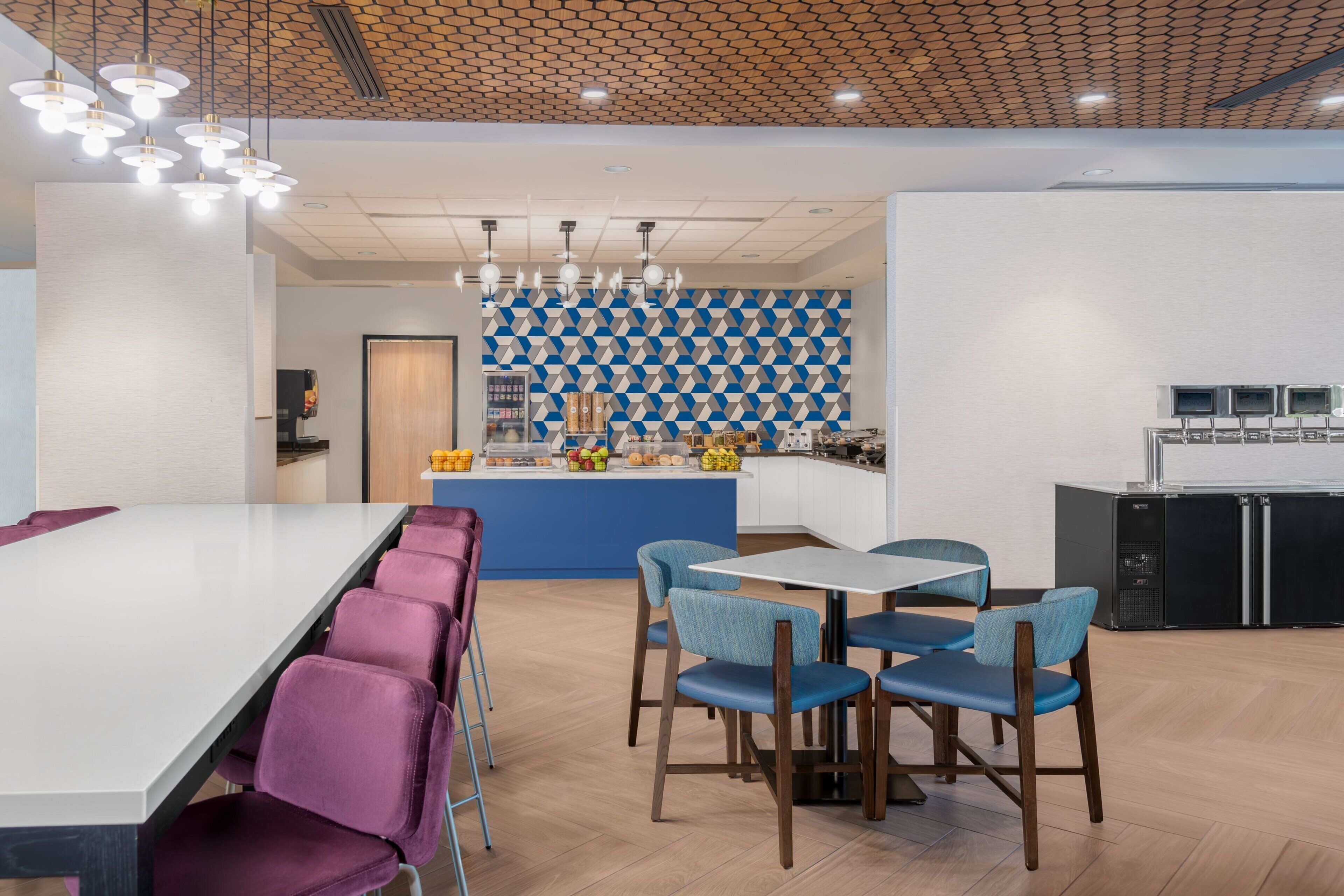 A modern kitchen with a blue and white geometric backsplash, a central island with fruit and snacks, and a seating area with purple and blue chairs.