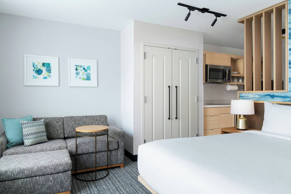 Una habitación de hotel moderna con un sofá gris, una mesita redonda de madera, una cama con colchón blanco, una kitchenette con microondas y un armario de dos puertas.